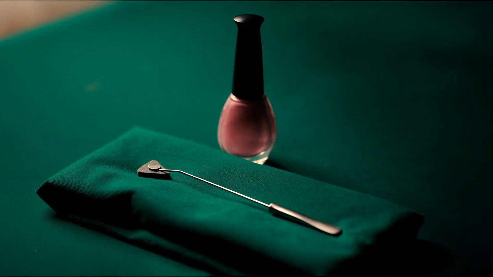 A steel cuticle pusher resting on folded green cloth beside a bottle of rose lacquer
